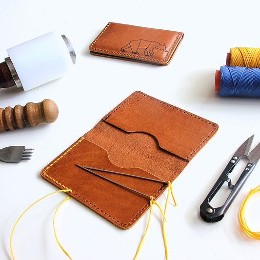 Close-up of a minimalist leather cardholder with hand-stitching. Tools and materials for leathercraft are arranged around it.