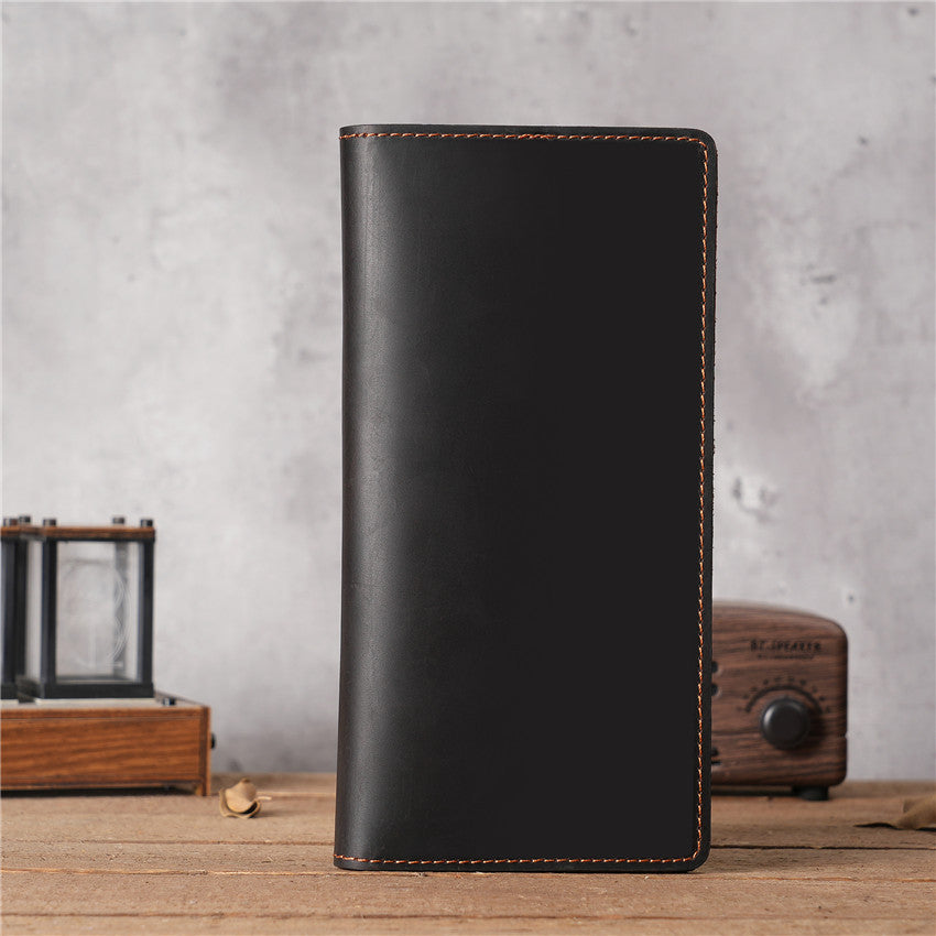 Dark Brown handmade oil waxed leather Long Snap Wallet, closed, standing upright on a rustic wooden surface with a blurred vintage radio and decorative glass box in the background.