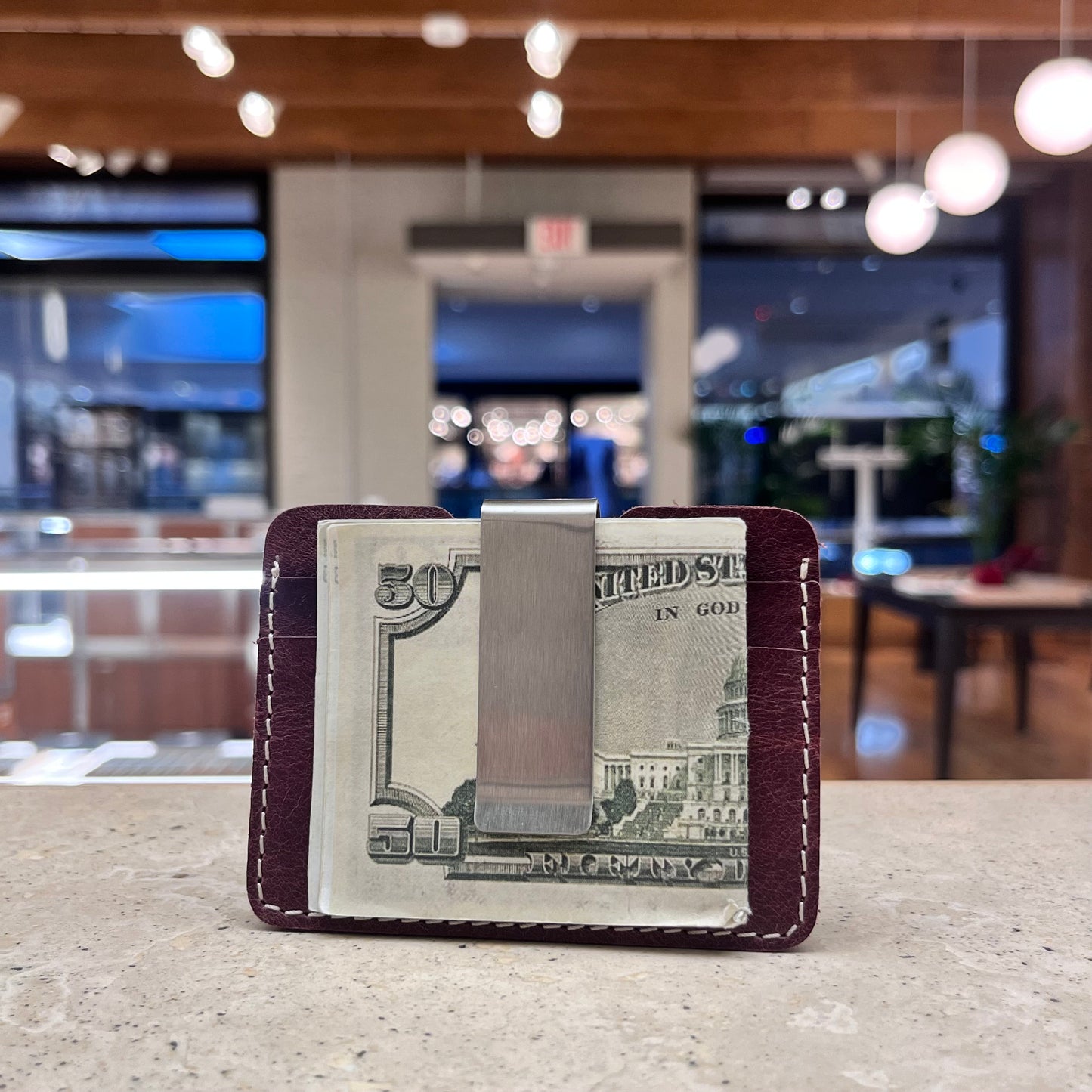 A compact burgundy leather card holder with a metal money clip securing US dollar bills.