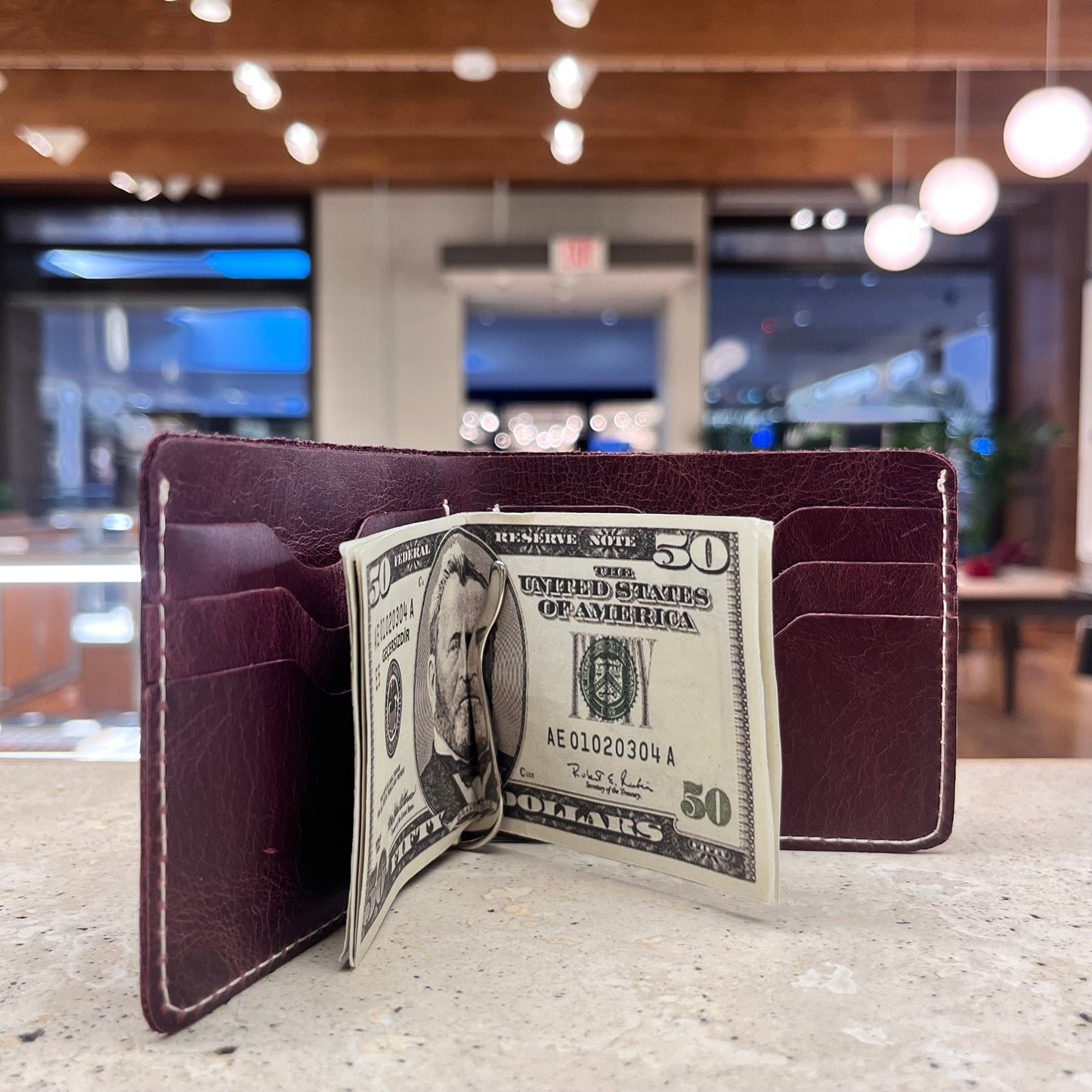 A slim burgundy leather bifold wallet with a money clip holding several US dollar bills.