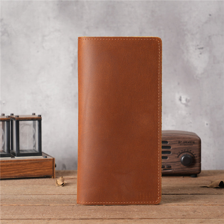 Tan brown handmade oil waxed leather Long Snap Wallet, closed, standing upright on a rustic wooden surface with a blurred vintage radio and decorative glass box in the background.