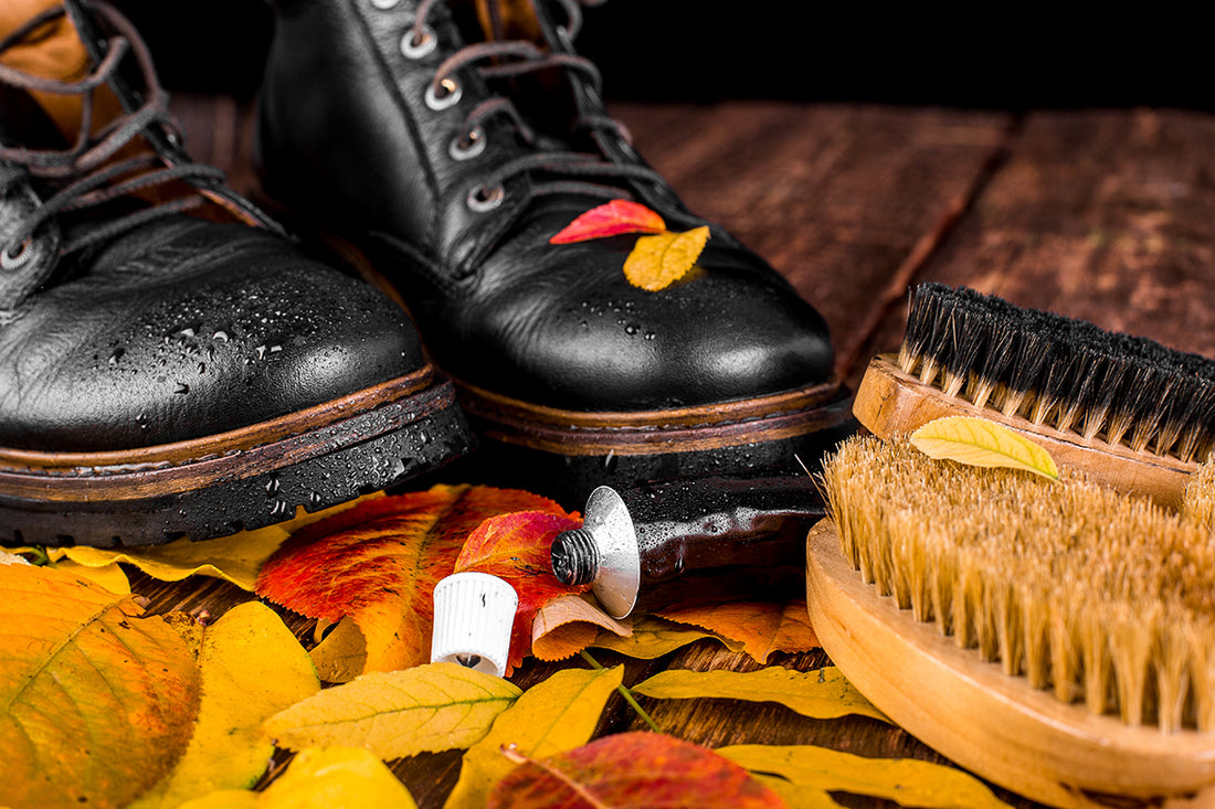 A pair of black leather boots with water droplets, a shoe polish applicator, a white cap, and two shoe brushes rest on a wooden surface surrounded by fallen autumn leaves.