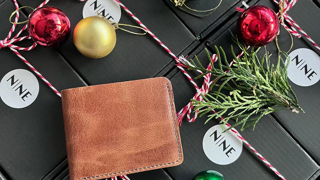 A festive flat lay of a NINE leather bifold wallet, surrounded by black gift boxes with NINE stickers, red and gold Christmas ornaments, and a sprig of evergreen.
