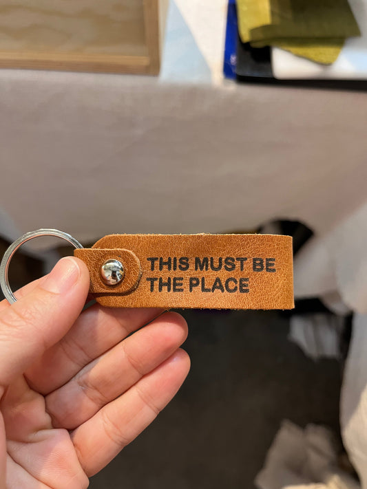 A person's hand holding a NINE Leather & Watch personalized tan brown leather keychain, showing a custom vertical engraving of the message "THIS MUST BE THE PLACE"