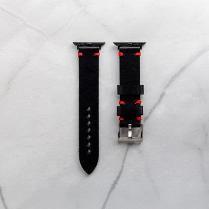 Handcrafted black full-grain leather watch band for Apple Watch featuring red artisan stitching and black adapters on a white marble background.
