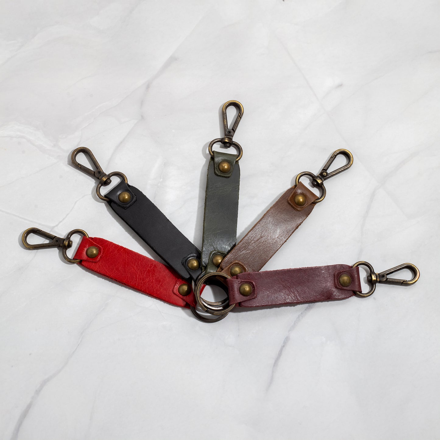 Five leather keychains fanned out in a circle showing available colors: Red, Black, Green, Brown, and Burgundy, all featuring antique brass swivel hooks and rivets.