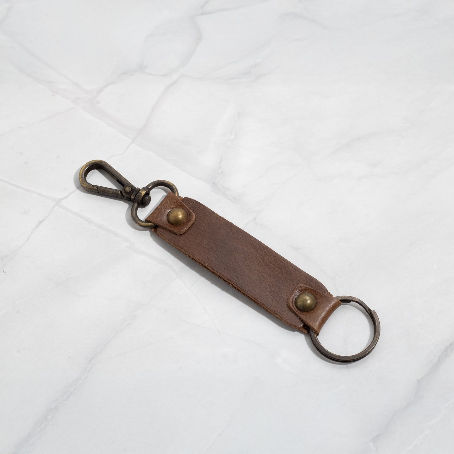 Brown leather keychain strap with vintage-style antique brass lobster clasp and key ring lying flat on a white marble surface.