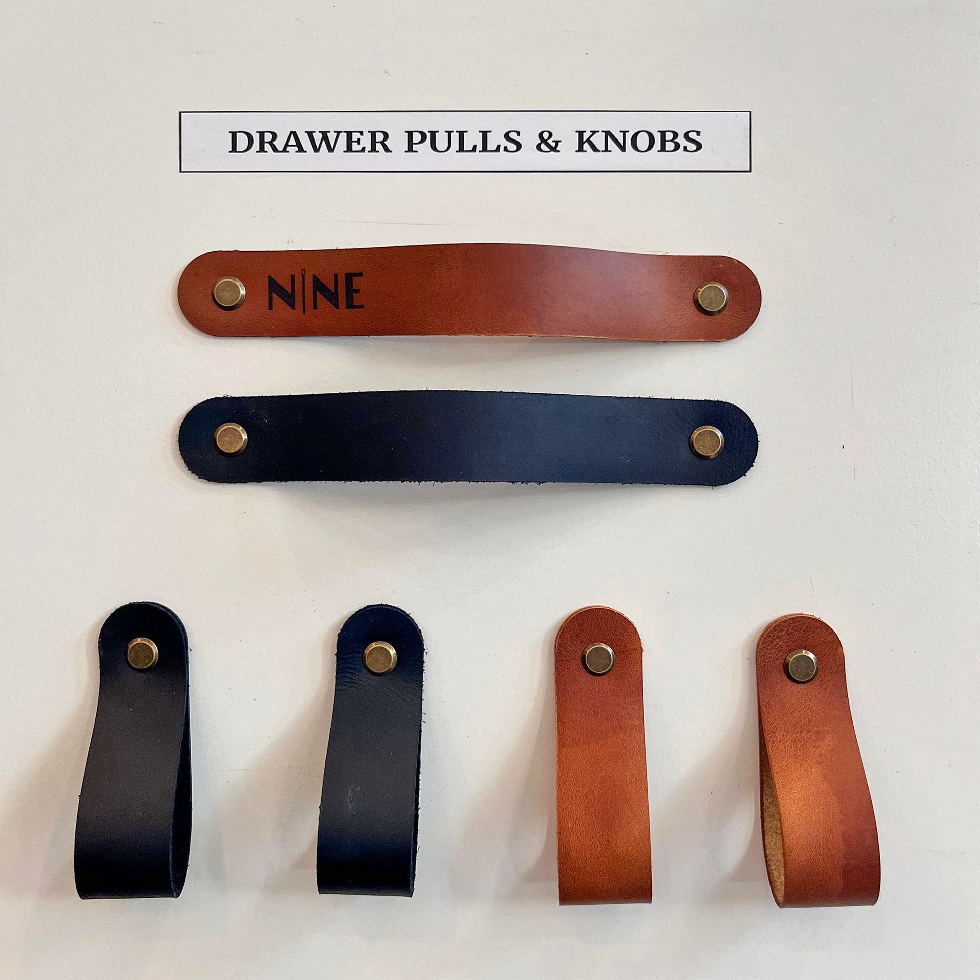 Frontal view of artisan leather drawer pulls and loop knobs in black and tan, featuring the NINE logo and brass fasteners.