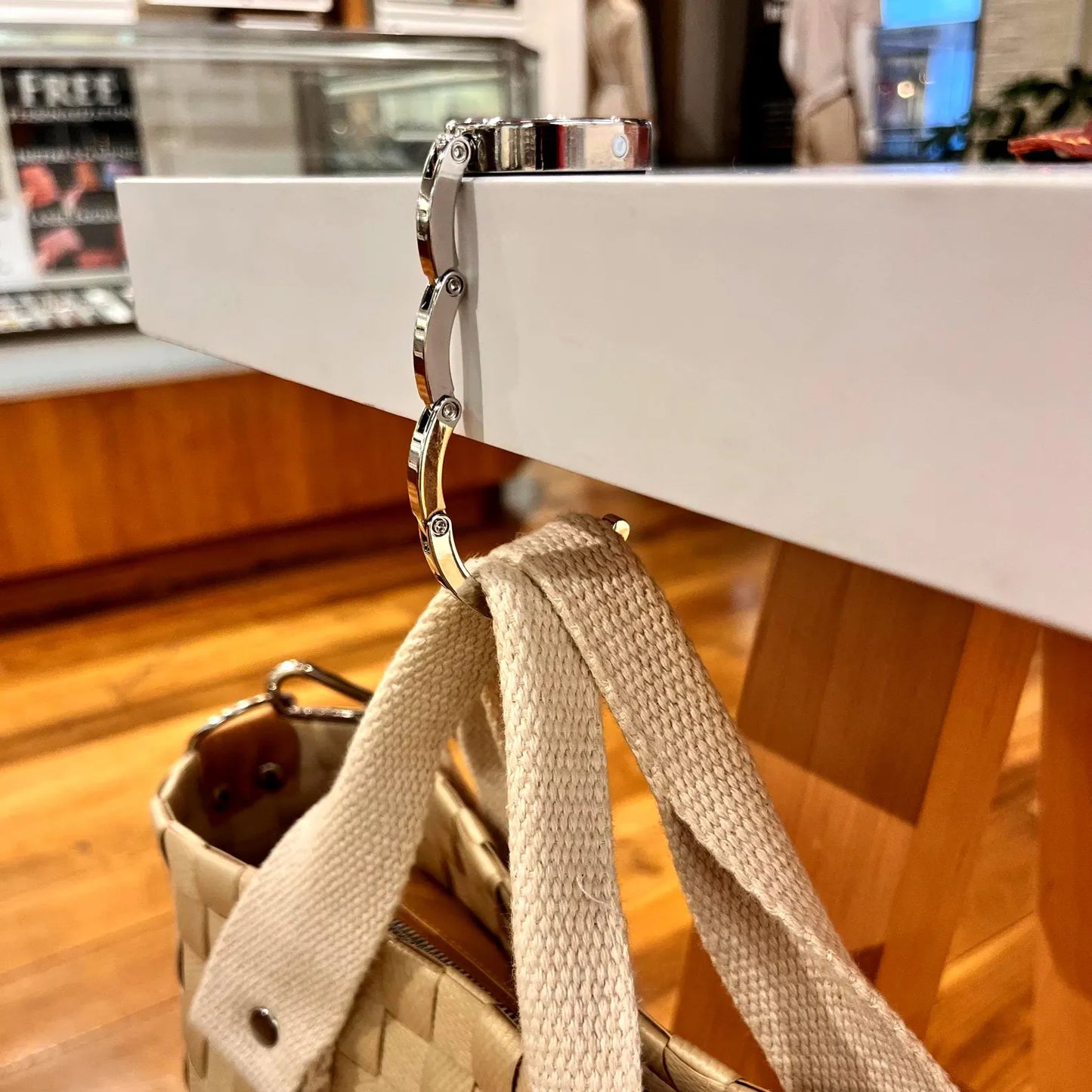 Close-up side view of the zinc alloy folding mechanism of a bag hanger extended over a table edge, supporting a beige tote bag.