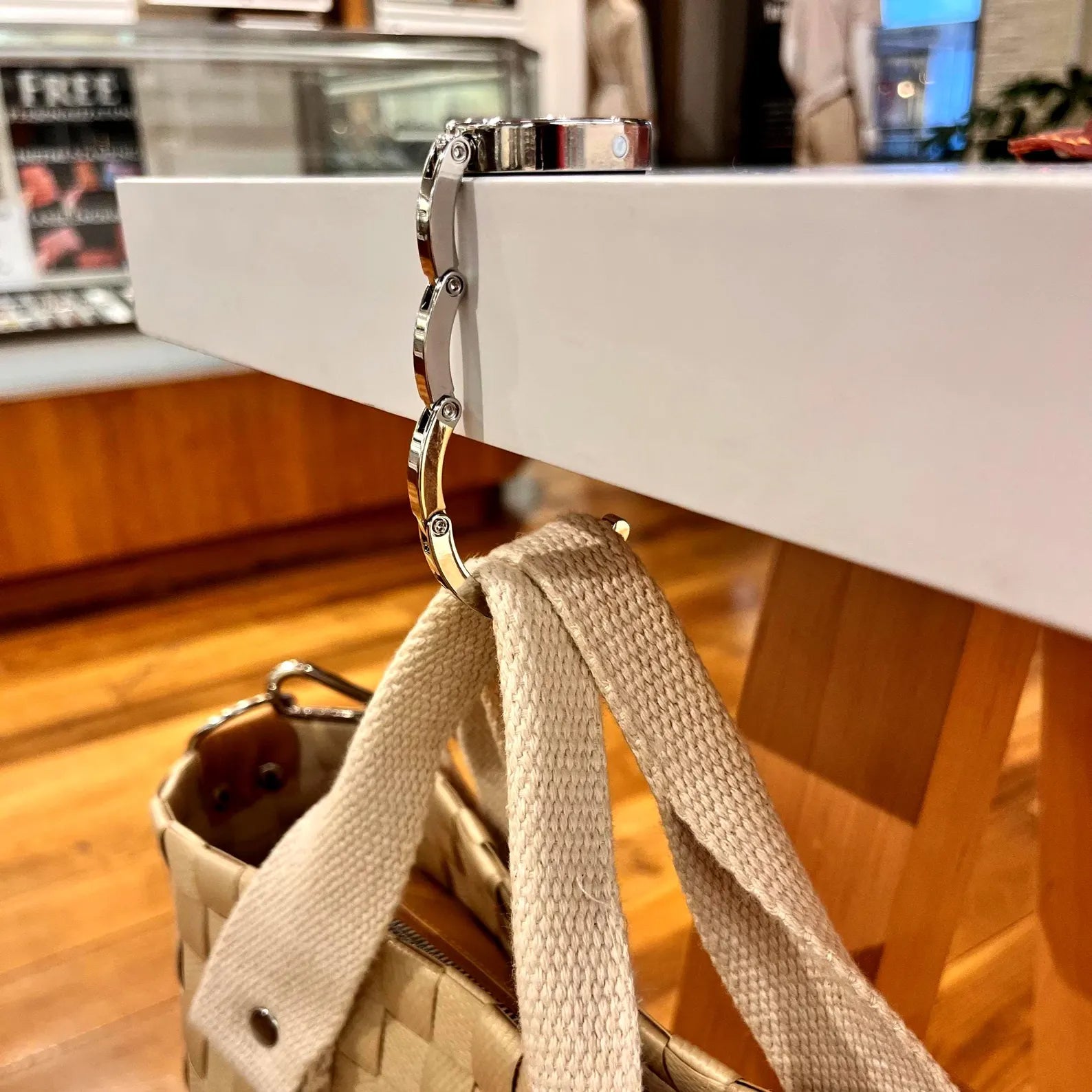 Close-up side view of the zinc alloy folding mechanism of a bag hanger extended over a table edge, supporting a beige tote bag.