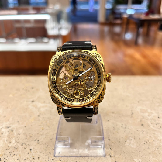 Customized NINE automatic skeleton watch, gold-colored engraved case, luminous display, and black handmade leather band with gold butterfly clasp, slight side angle view showing case detail.