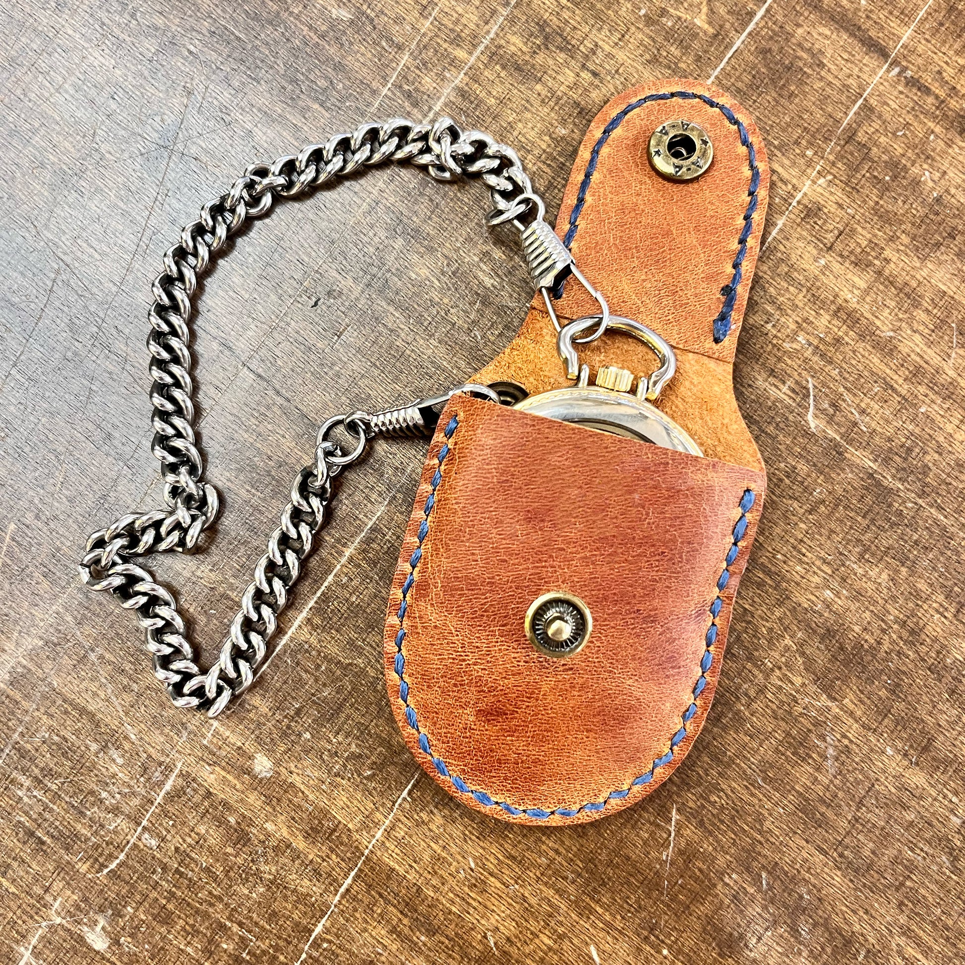 Open tan leather pocket watch sleeve with contrasting blue hand-stitching, showing a silver pocket watch sliding into the snug, custom-fit holder with a chain attached.