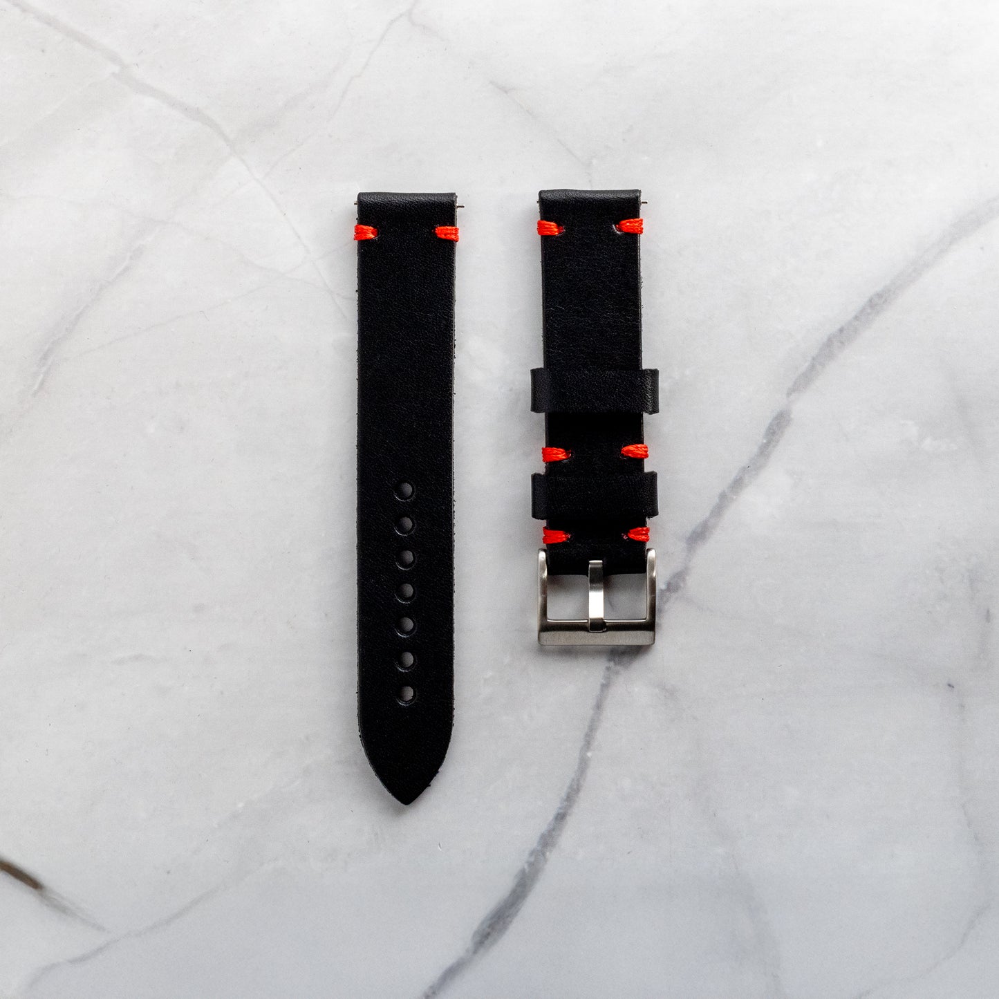 Top-down view of a handmade black full-grain leather watch band featuring vibrant red artisan stitching and a classic silver buckle on a marble background.