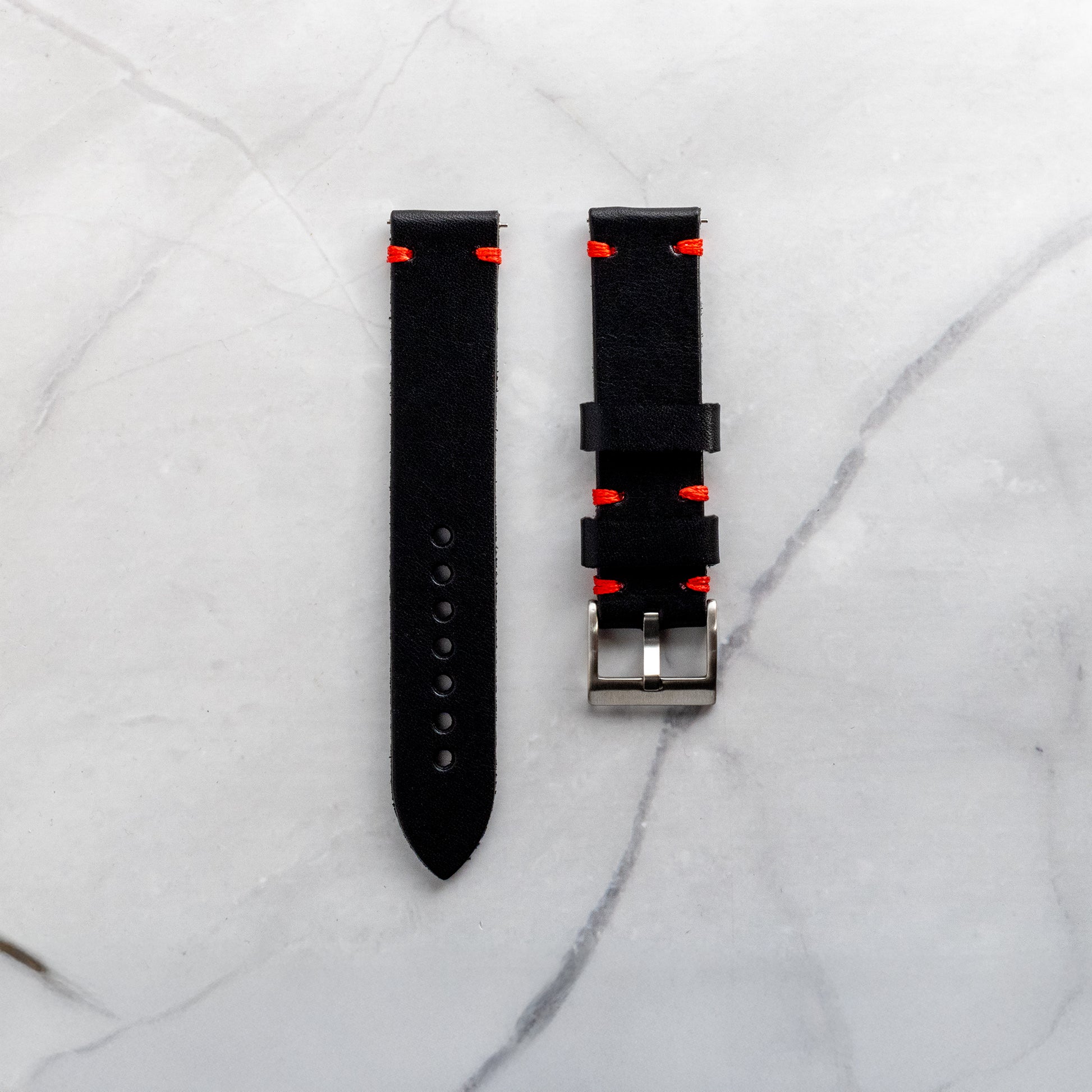 Top-down view of a handmade black full-grain leather watch band featuring vibrant red artisan stitching and a classic silver buckle on a marble background.