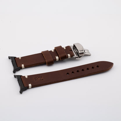 A brown leather watch strap with silver-toned metal hardware and white stitching, displayed on a clean white background. One end features a black plastic connector, and the other is shown with the buckle open.