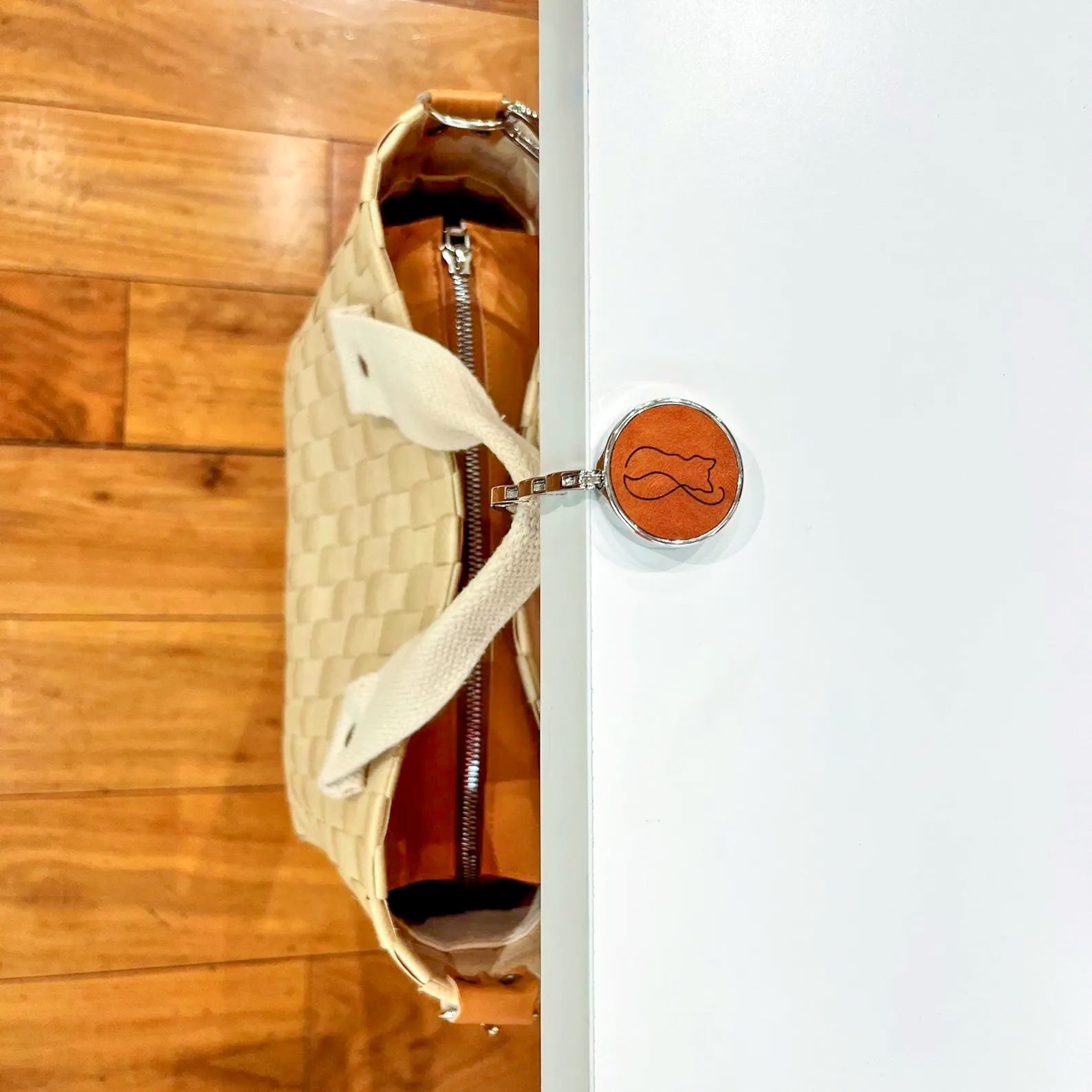 Tan leather bag hanger with a laser-engraved cat outline design, securely holding a woven handbag on the edge of a white table.