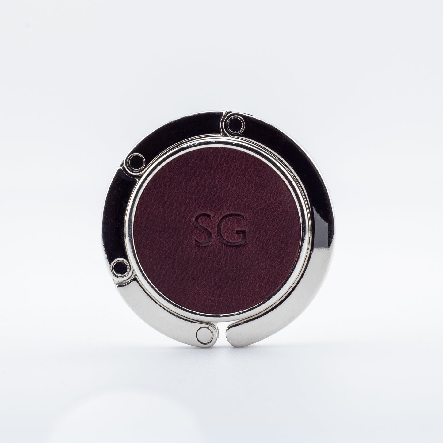 Close-up of a polished metal foldable purse hanger featuring a burgundy full-grain leather insert, custom monogrammed with the initials SG.