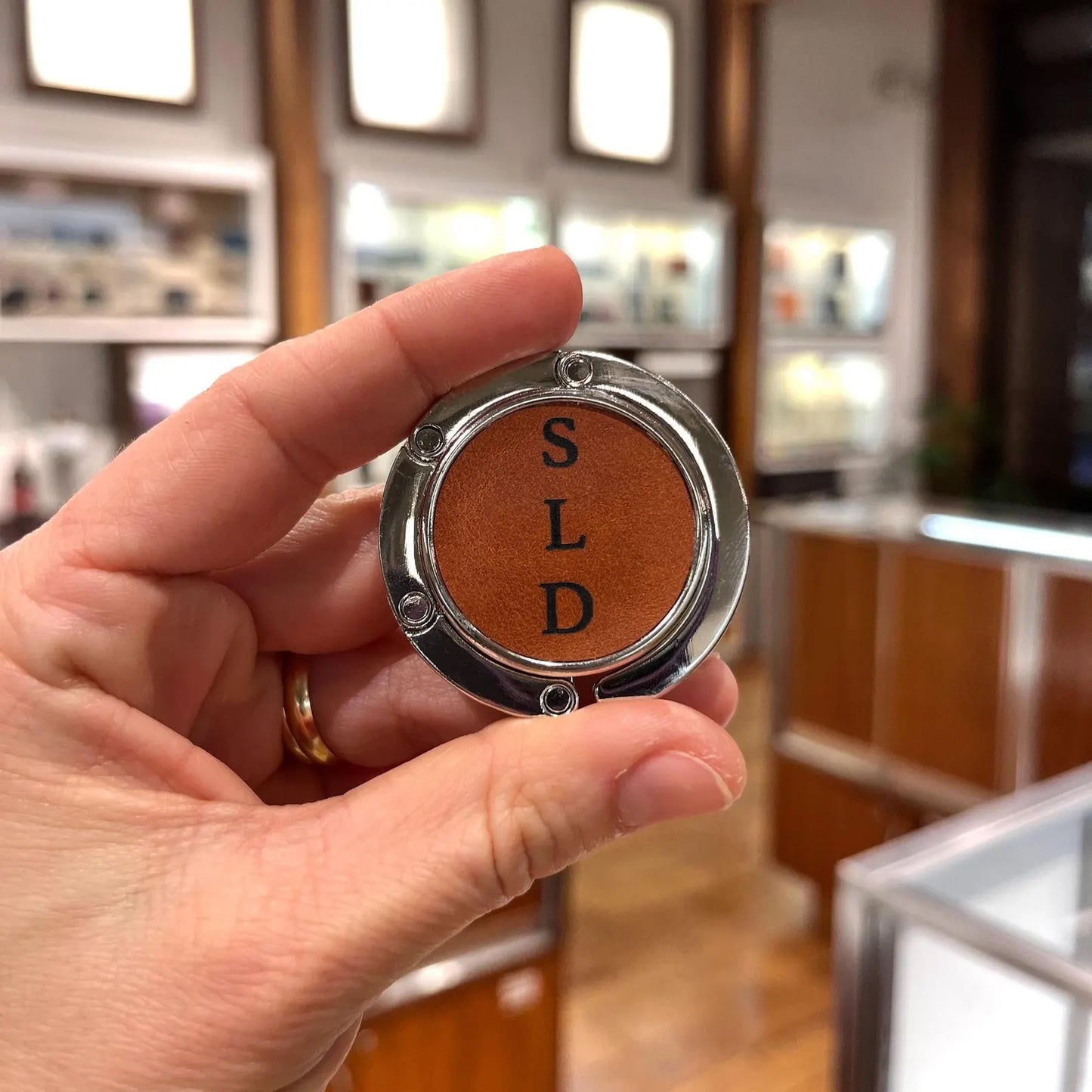 Hand holding a folded custom bag hanger featuring tan leather with "SLD" initials laser engraved in a vertical format.