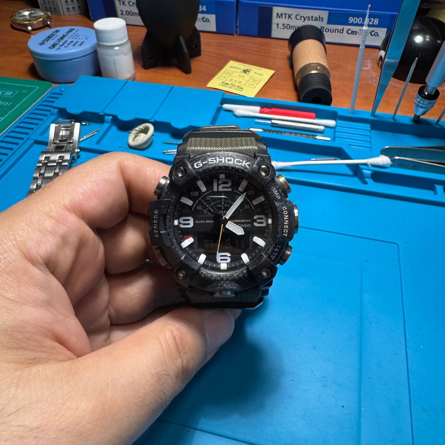 Front view of a restored Casio G-Shock Mudmaster GG-B100 watch featuring a black carbon bezel and olive green strap, held over a blue watchmaker's mat.