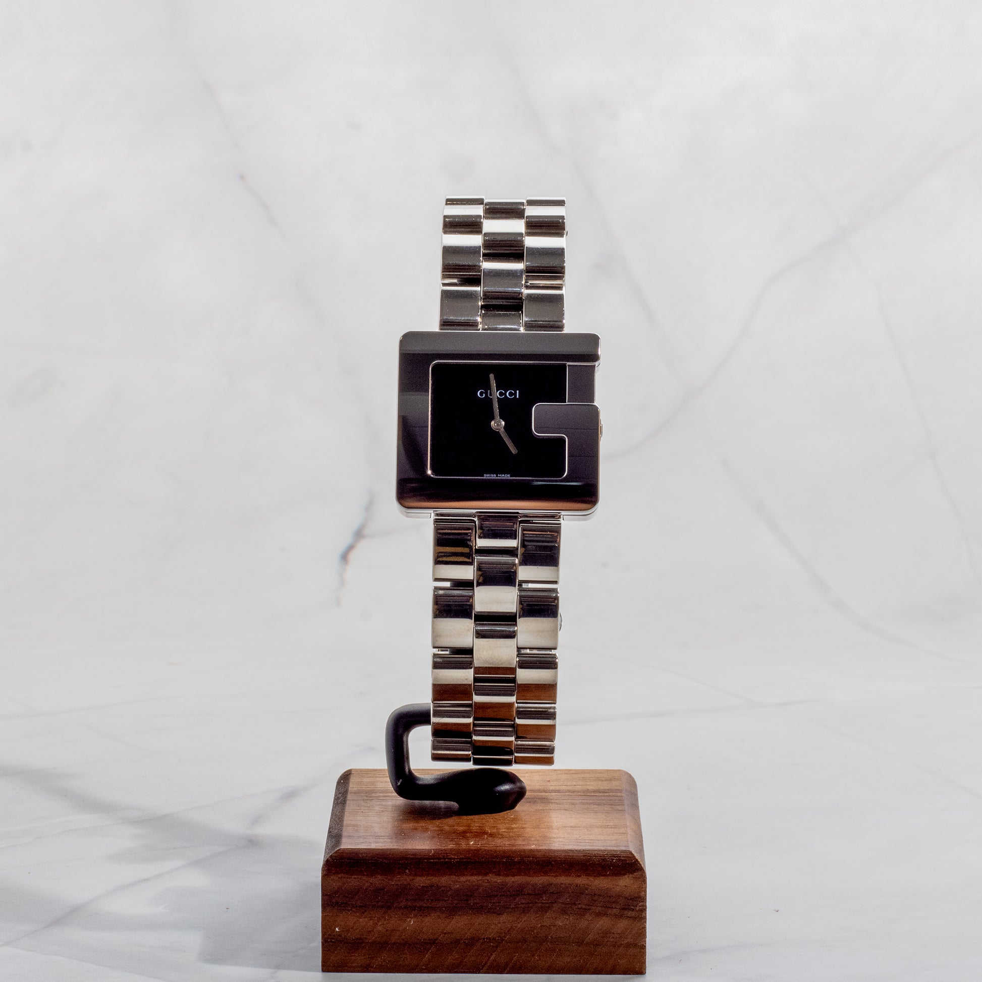 A professionally restored Gucci 3600M stainless steel watch with the iconic "G" shape case and a minimalist black dial, displayed on a wooden stand against a marble background.
