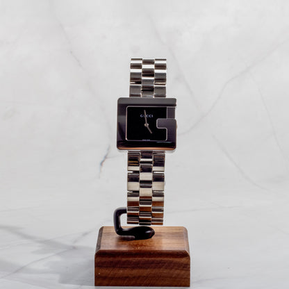 A professionally restored Gucci 3600M stainless steel watch with the iconic "G" shape case and a minimalist black dial, displayed on a wooden stand against a marble background.