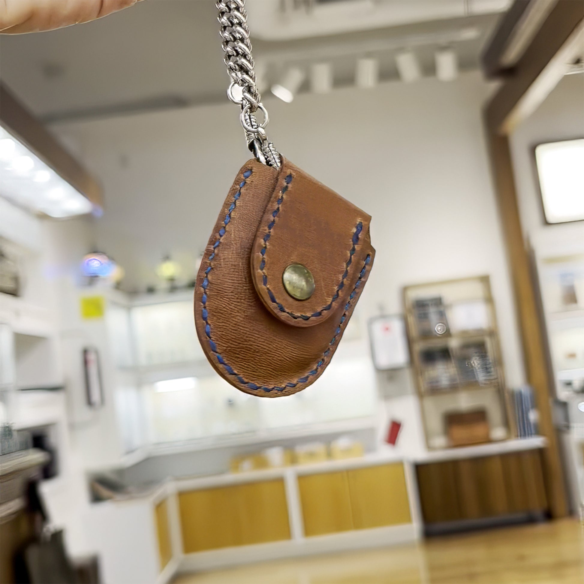 Tan leather pocket watch case hanging securely by a metal watch chain, demonstrating the heavy-duty snap closure and durable saddle-stitched edges.