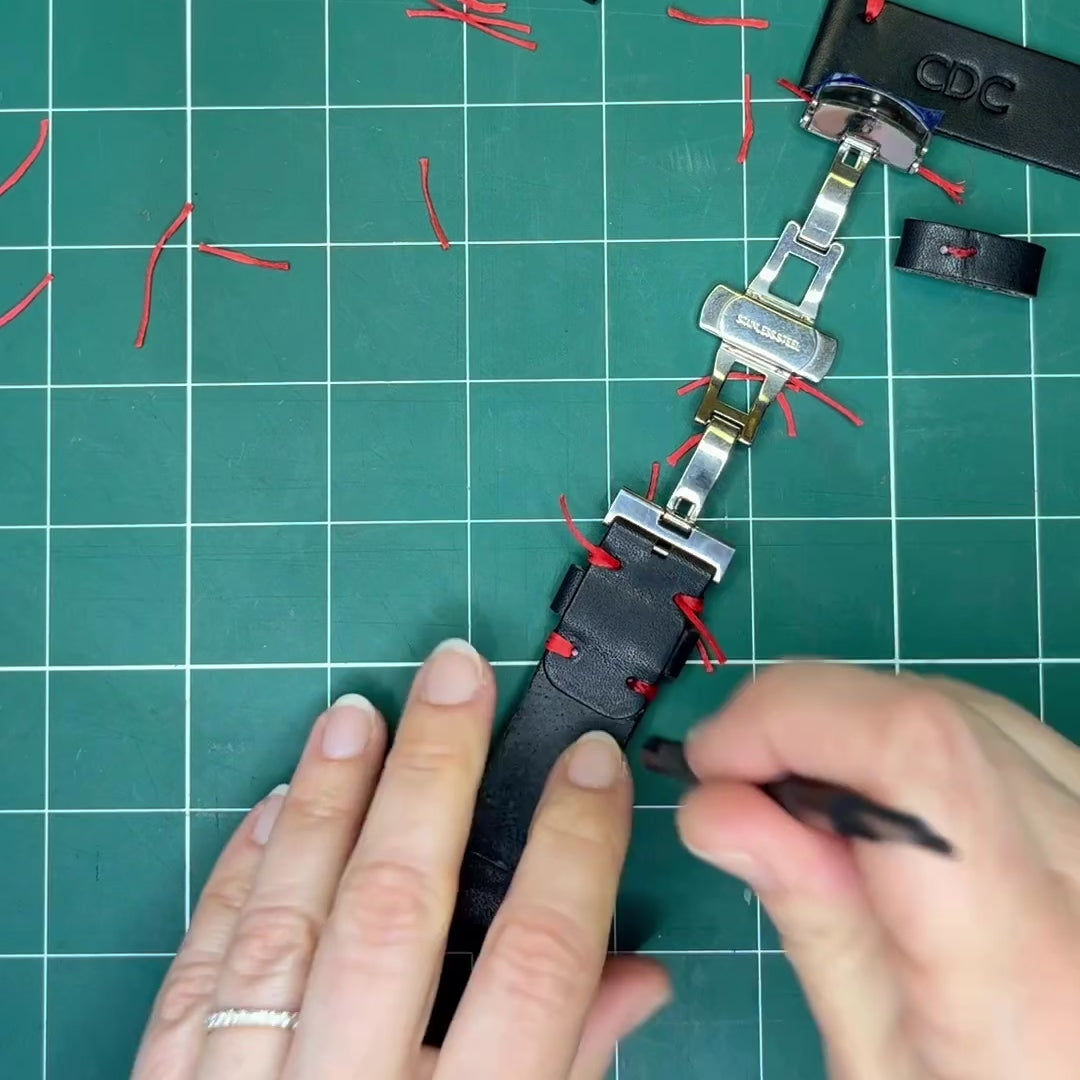 A craftsman using precision tools and heat to seal the custom red thread stitching on a handmade black full-grain leather watch band.